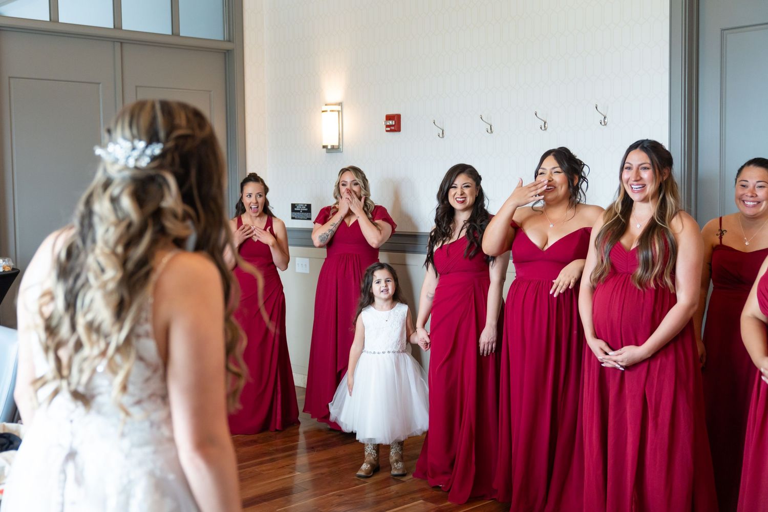 Bridesmaids in red dresses line up against a white wall during wedding preparations.