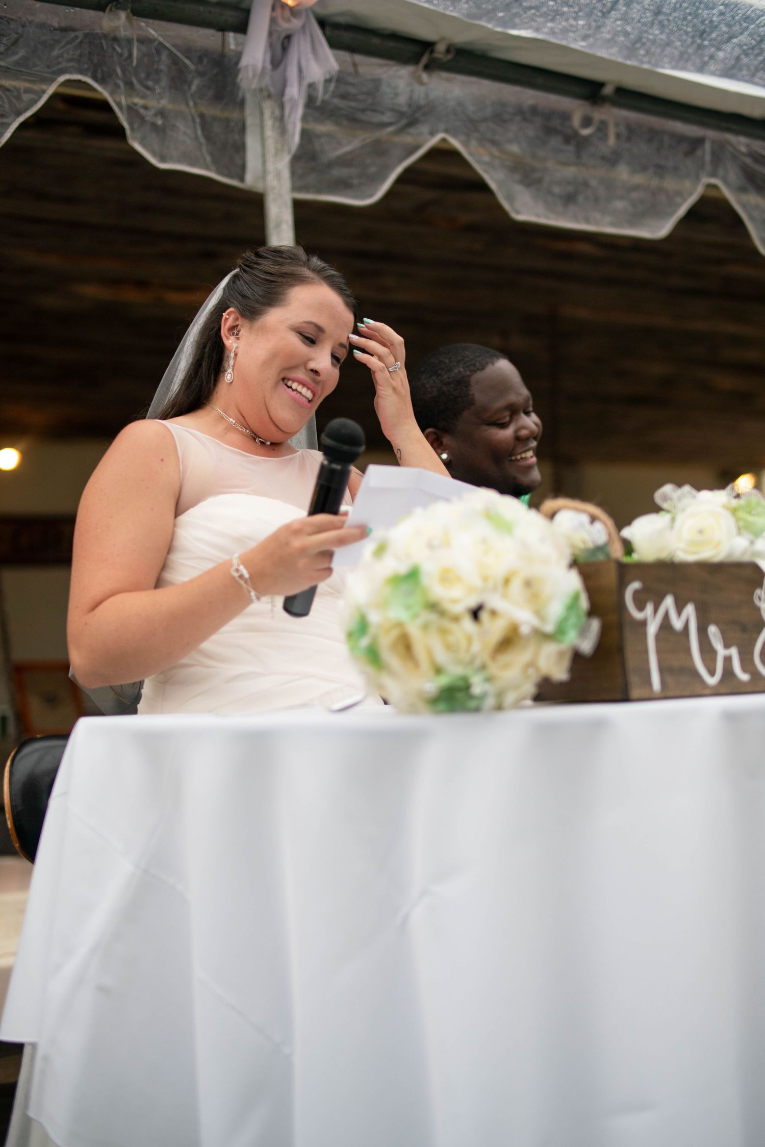 Wedding reception table setup with white linens and floral arrangements under a covered venue.