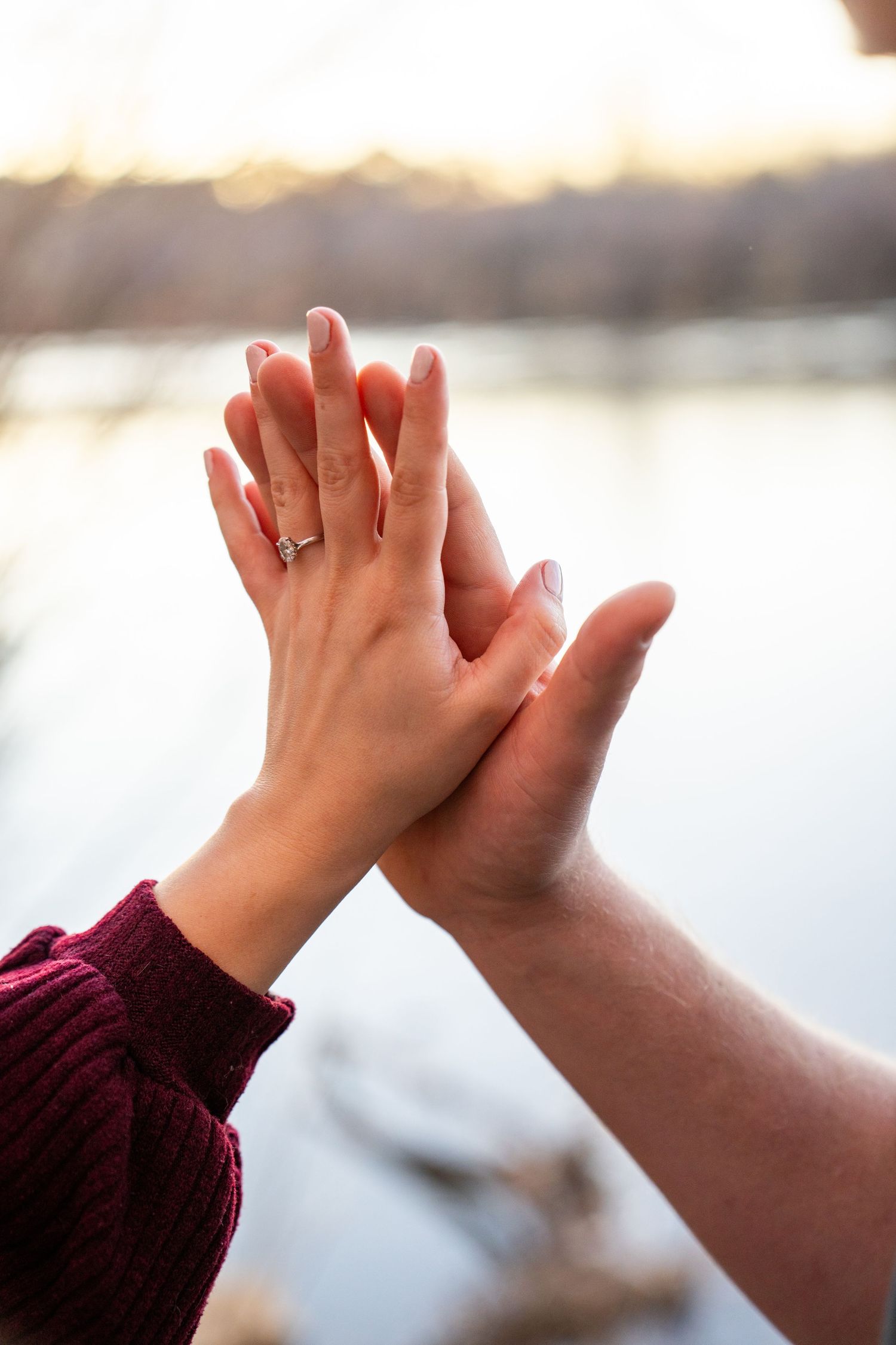 Two hands touching with an engagement ring on the woman's finger, set against a blurred lake backdrop at sunset.