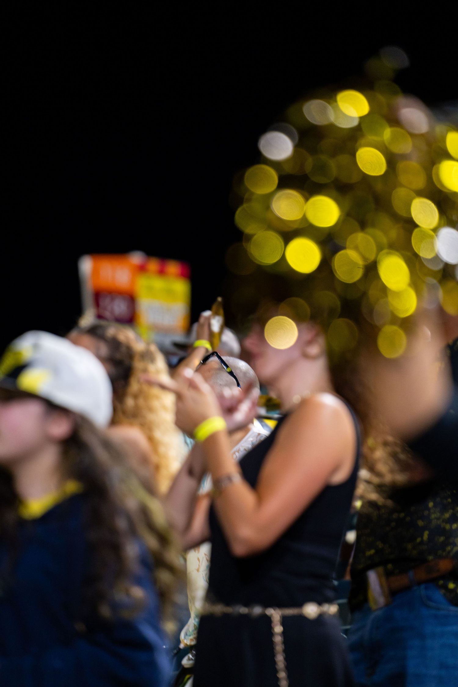 Blurred crowd at night event with golden bokeh lights and people in navy blue outfits holding pom-poms.
