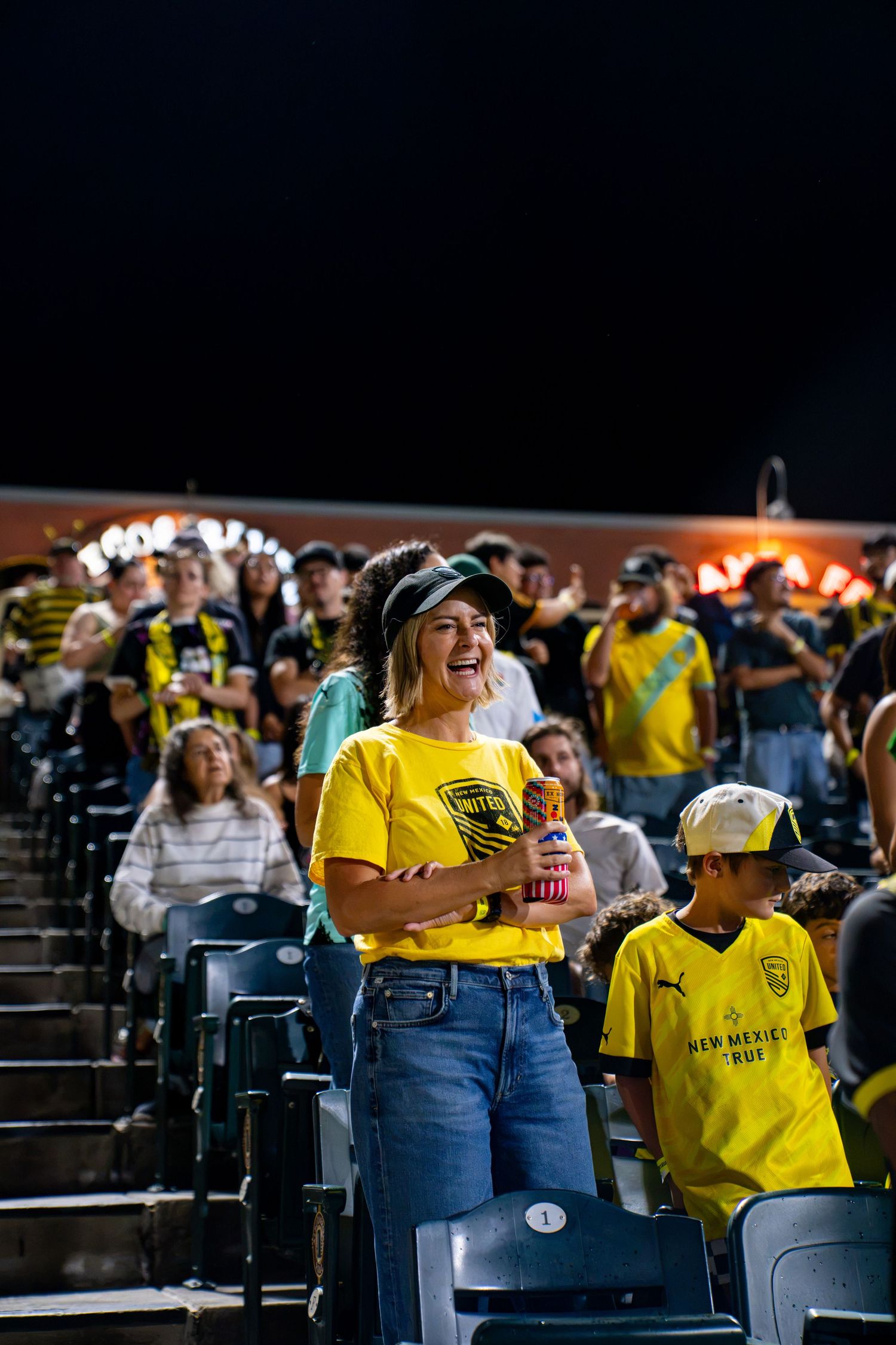 Excited female New Mexico United fan in yellow shirt laughs in stadium stands during night soccer match.