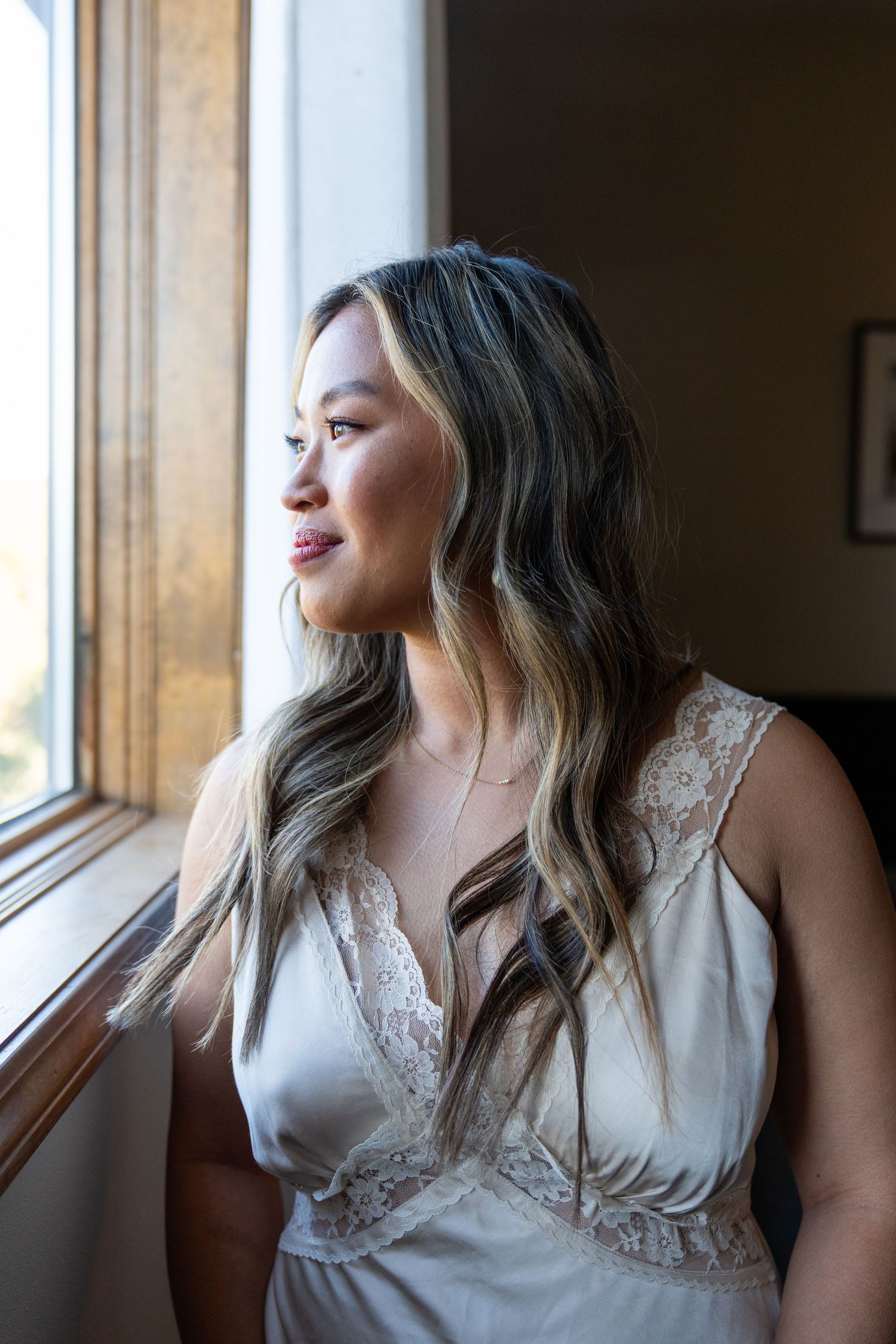 Soft natural window light illuminates someone in elegant attire looking outside.