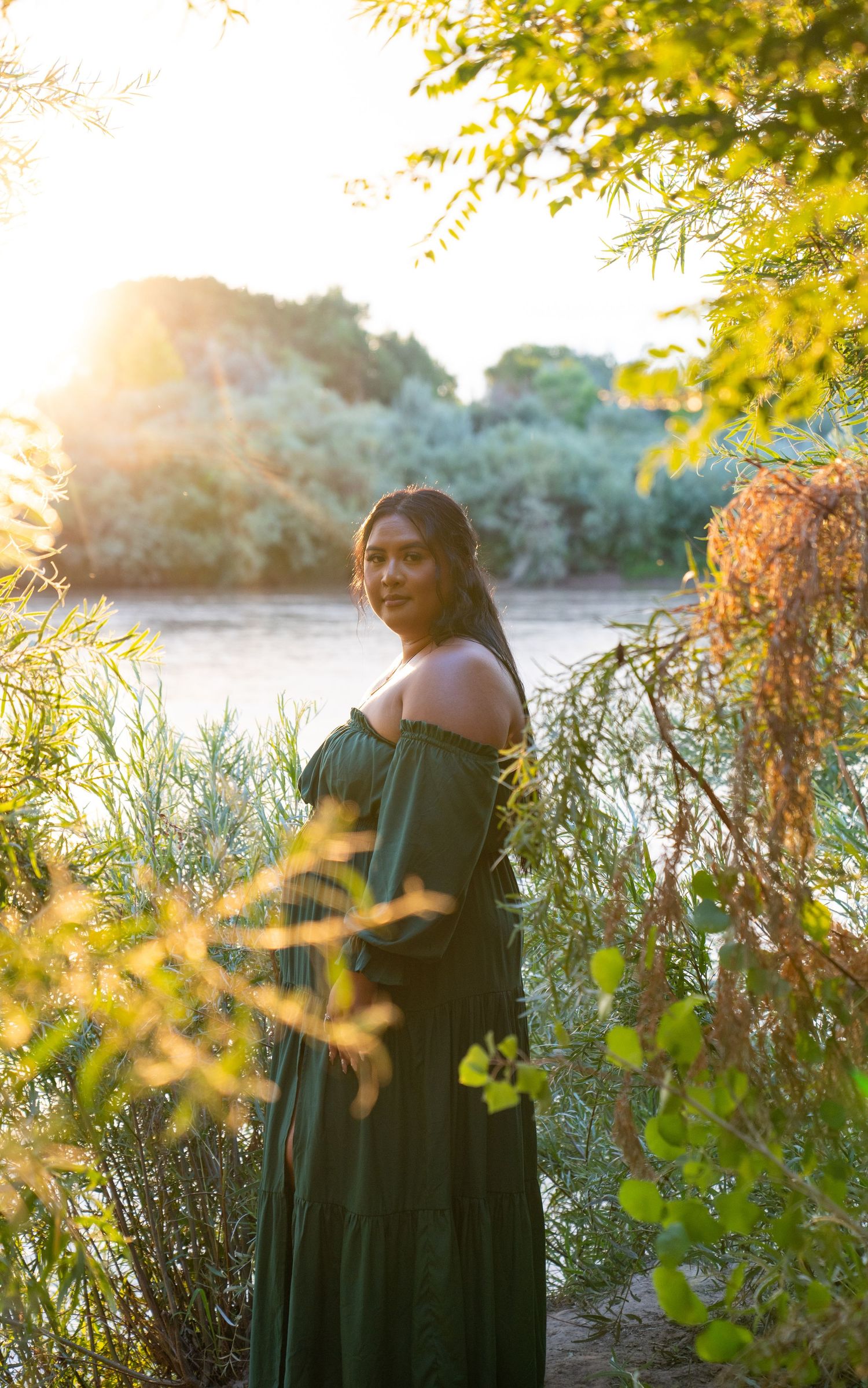 Sunset portrait in green dress by waterside with golden light filtering through foliage.