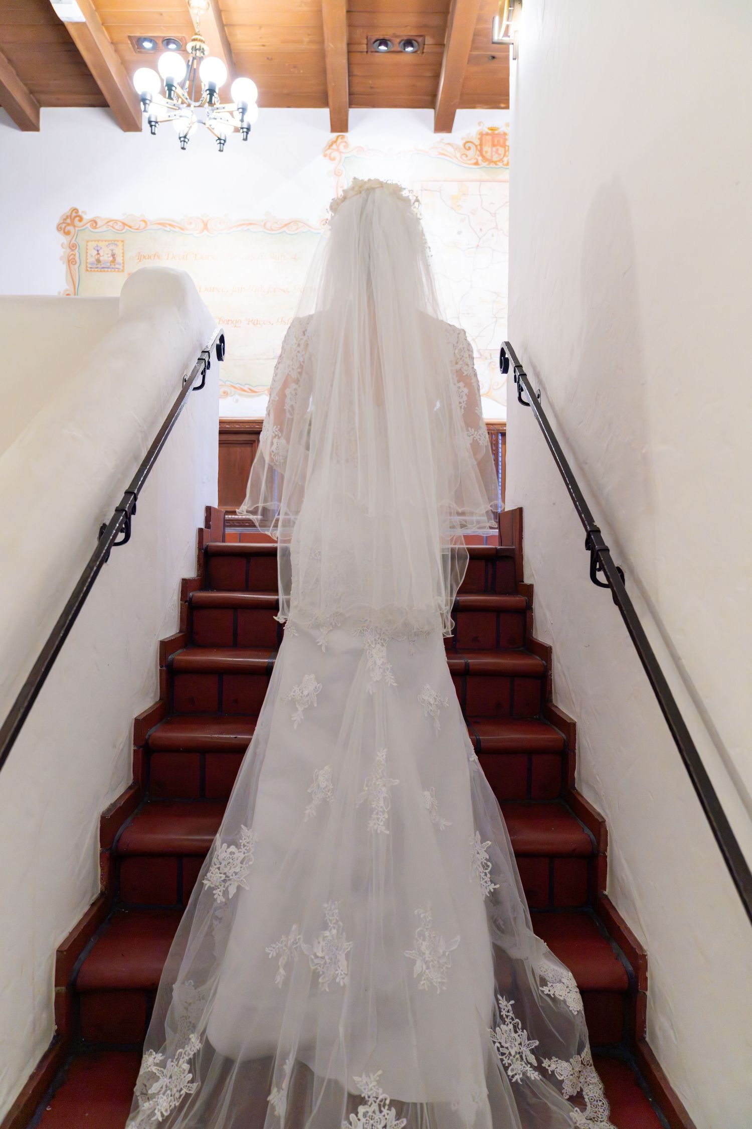 Bride in lace wedding gown and long veil ascending red-carpeted stairs in elegant venue.