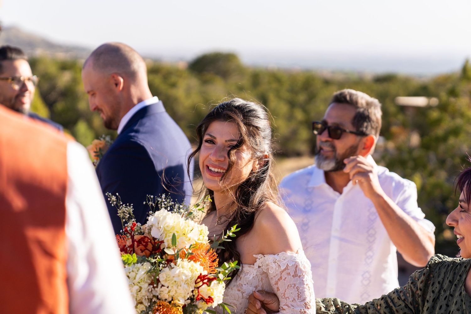 Smiling bride holding colorful bouquet at outdoor wedding ceremony with guests in background.