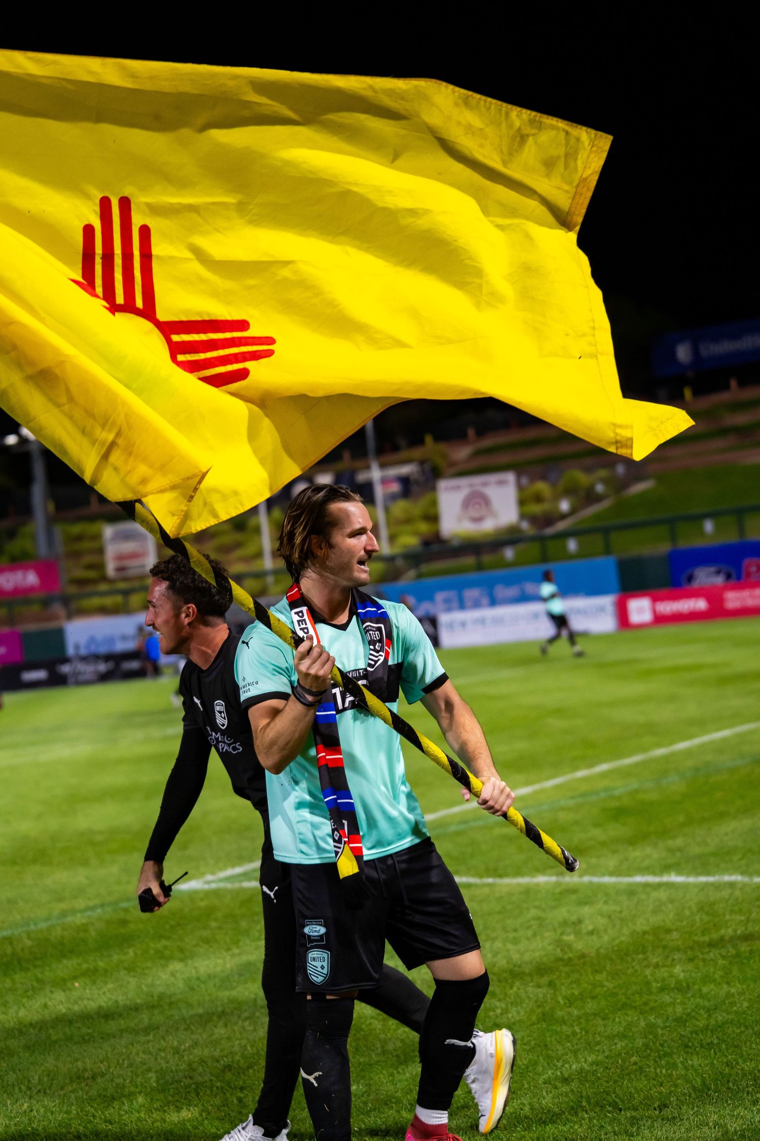 Soccer players wave a yellow New Mexico state flag on a green stadium field during a night game.