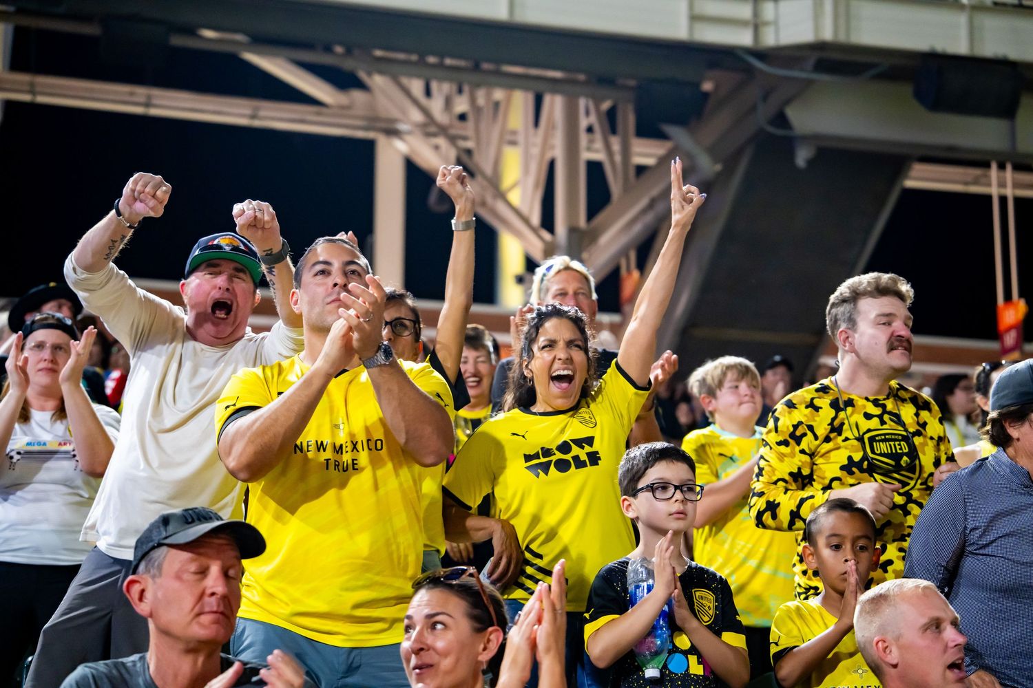 Excited fans in yellow shirts cheering at a sports event, raising fists and clapping in a crowded stadium.