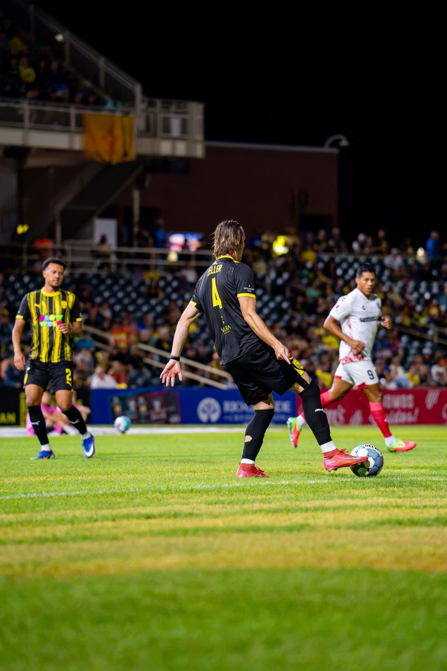 Soccer player number 4 in yellow controls the ball during a night match with teammates and opponents on a green field.