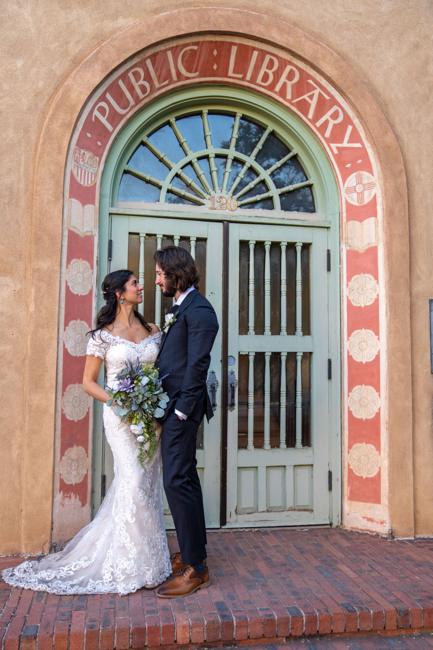 A wedding couple shares intimate moments in front of a historic library entrance with arched doorways and brick details.