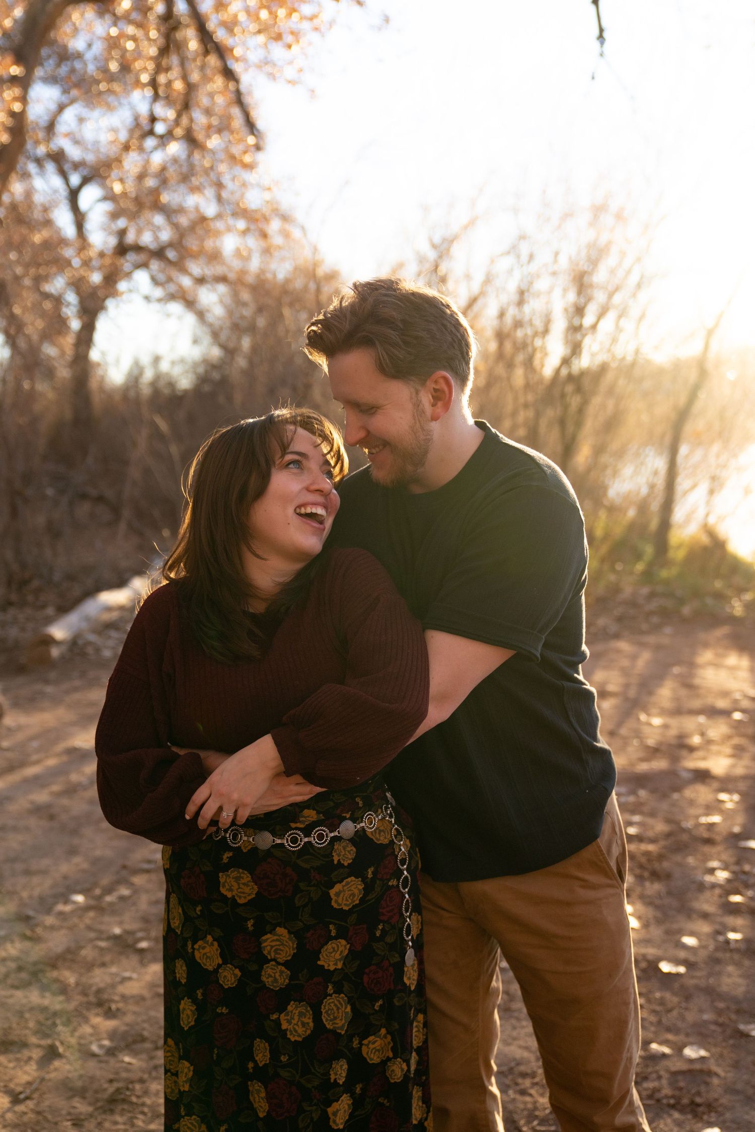 A happy couple embraces outdoors at golden hour, laughing together on a sunlit dirt path surrounded by autumn trees.