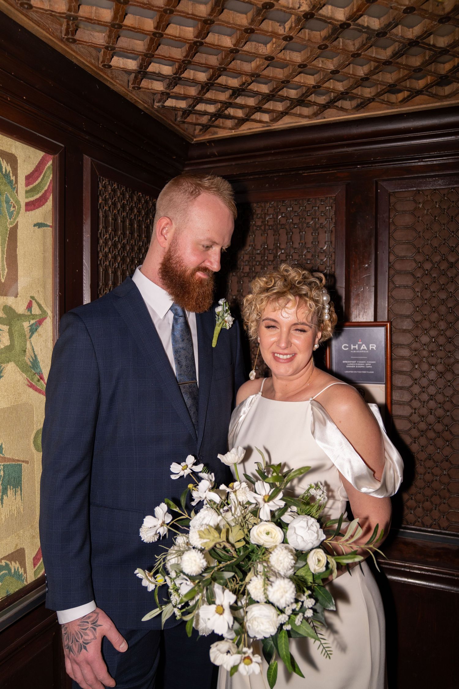 Bride and groom in elegant venue, groom in navy suit with beard, bride holding white floral bouquet.