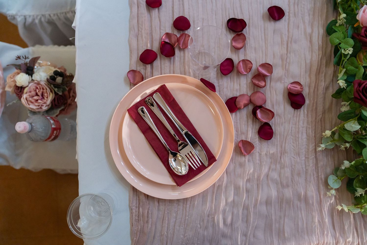 Pink place setting with silverware and rose petals on a wooden table at a wedding reception.