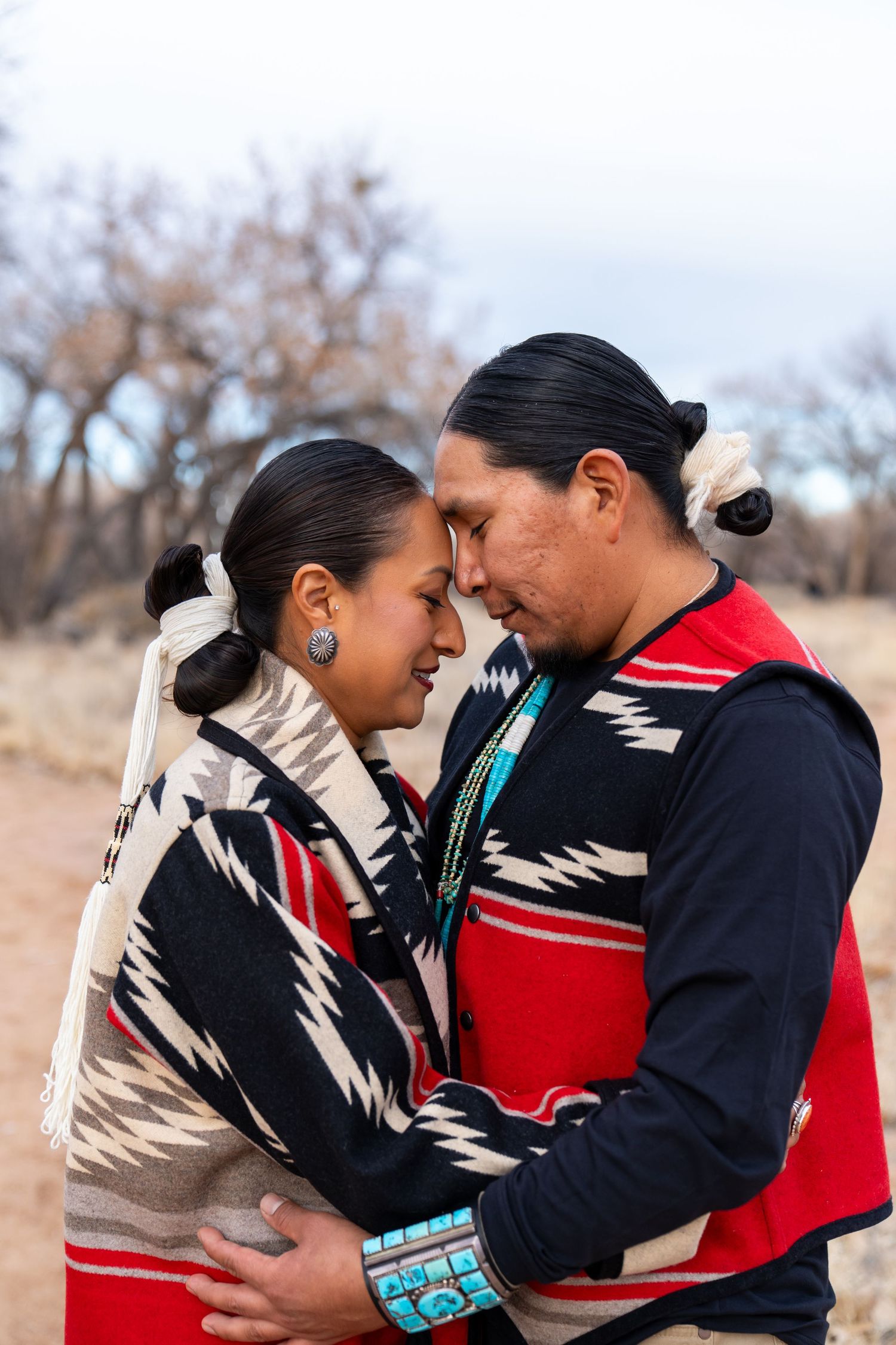 Two people in traditional Native American clothing embracing outdoors.