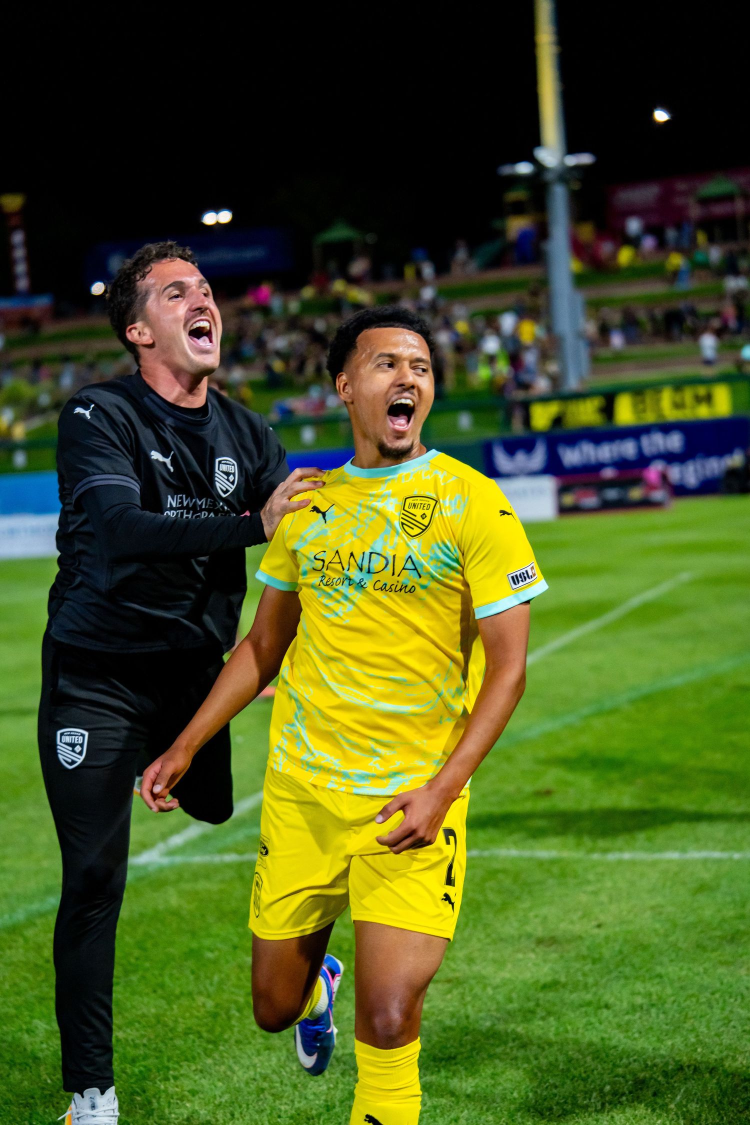 Two soccer players in yellow Sandia jerseys celebrate joyfully on the field during a night game.