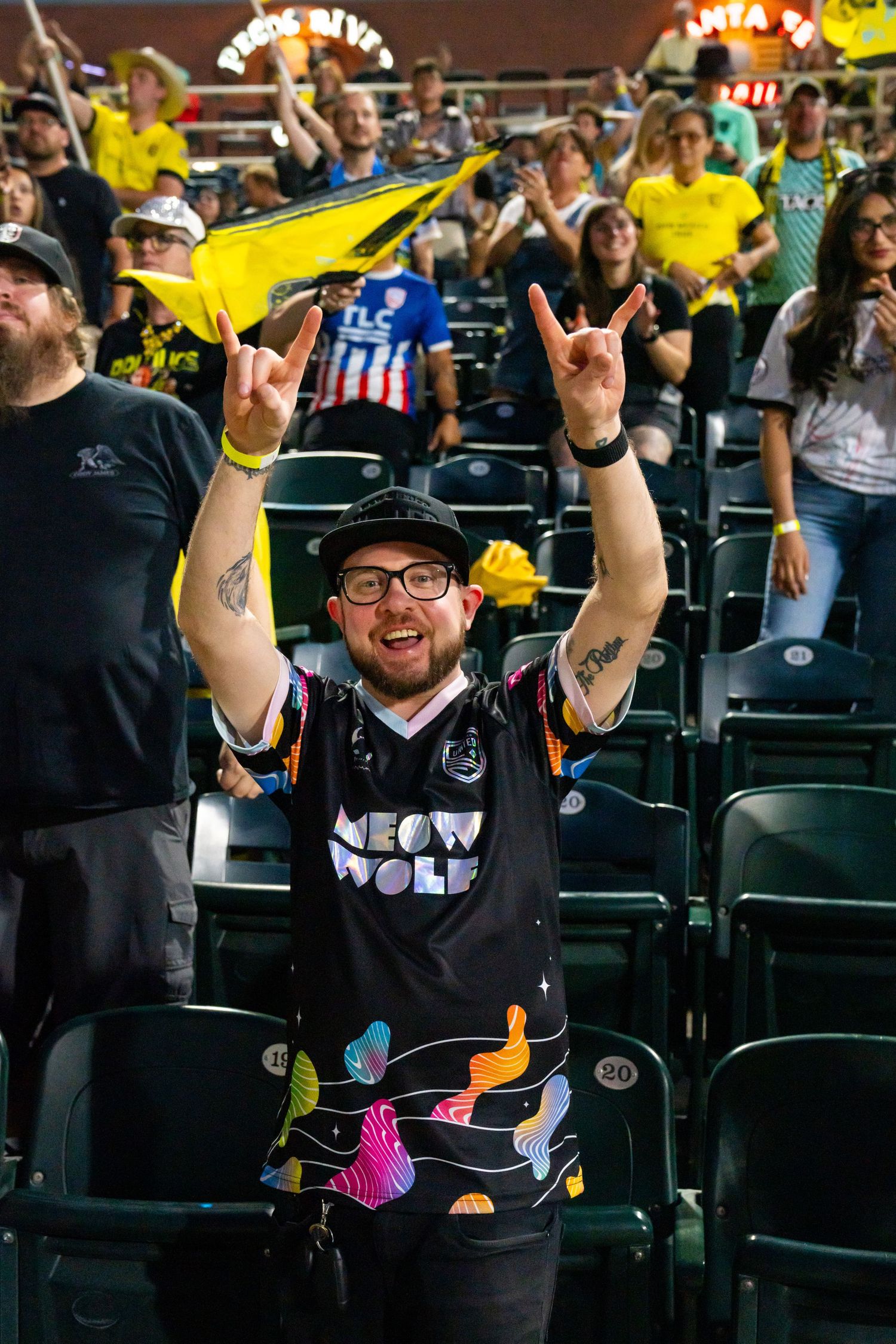 Excited Meow Wolf jersey fan makes rock on hand gestures at New Mexico United soccer match.