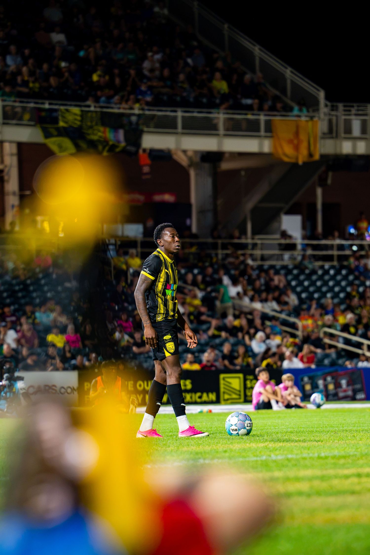 Soccer player in black and yellow kit stands near the ball during a night match at a packed stadium.