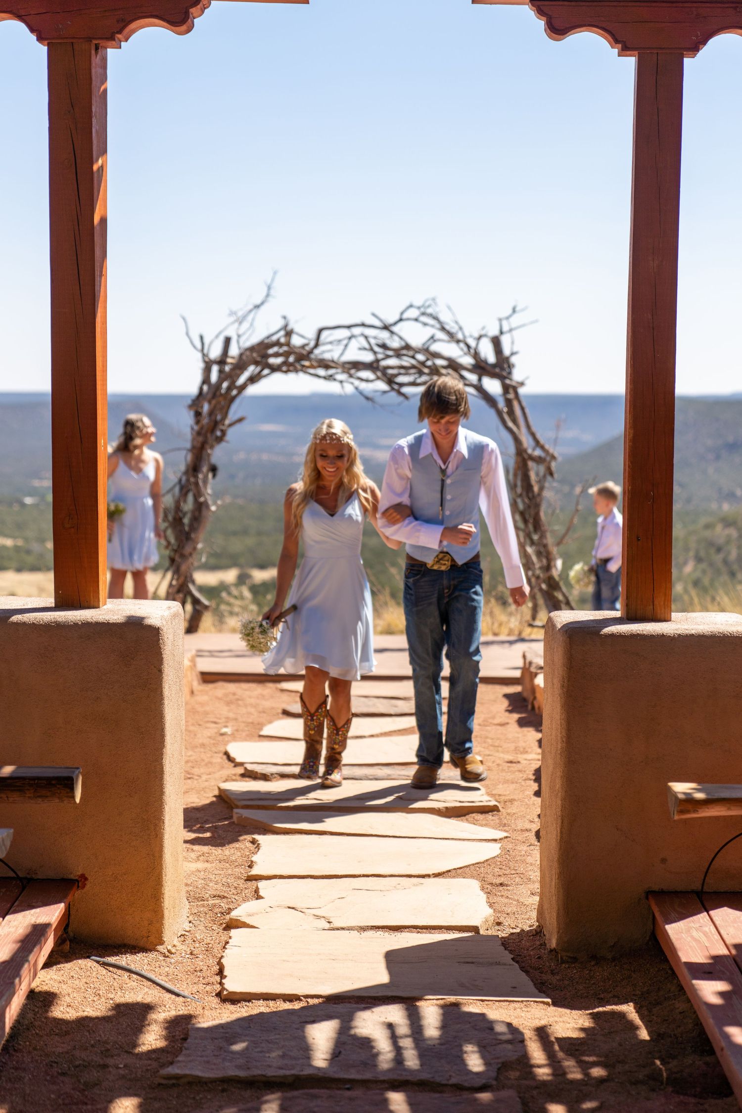 A couple walks through a rustic wooden archway with mountain views at an outdoor wedding venue in the desert.