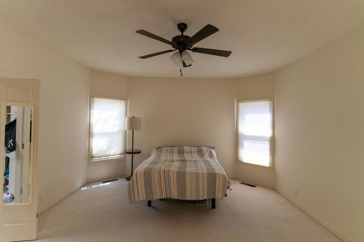 A bright, empty room with beige walls, ceiling fan, and multiple windows letting in natural light.