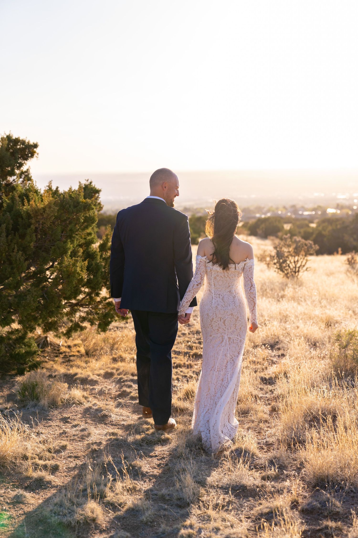 Bride in lace gown and groom in dark suit walk hand-in-hand through golden fields at sunset.