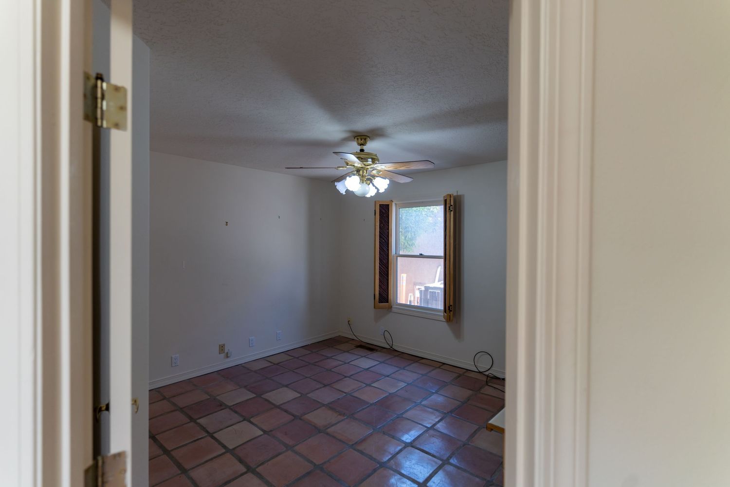 Empty bedroom interior with white walls, ceiling fan, and carpeted floor showing natural light from window.