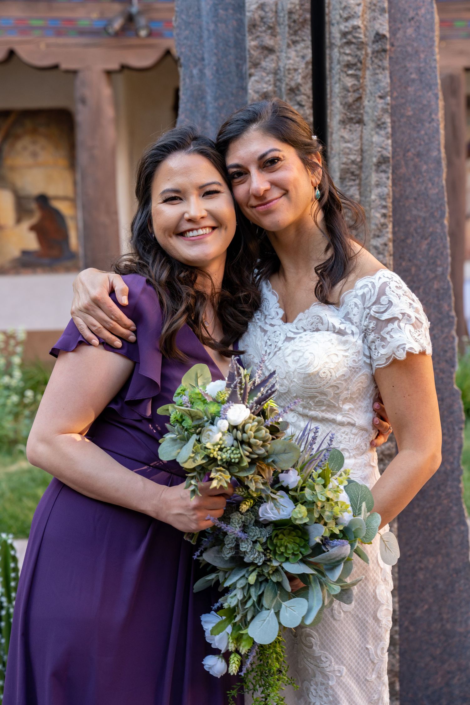 Two people embrace at a wedding, with one wearing a purple bridesmaid dress and the other in a white lace gown holding a bouquet.