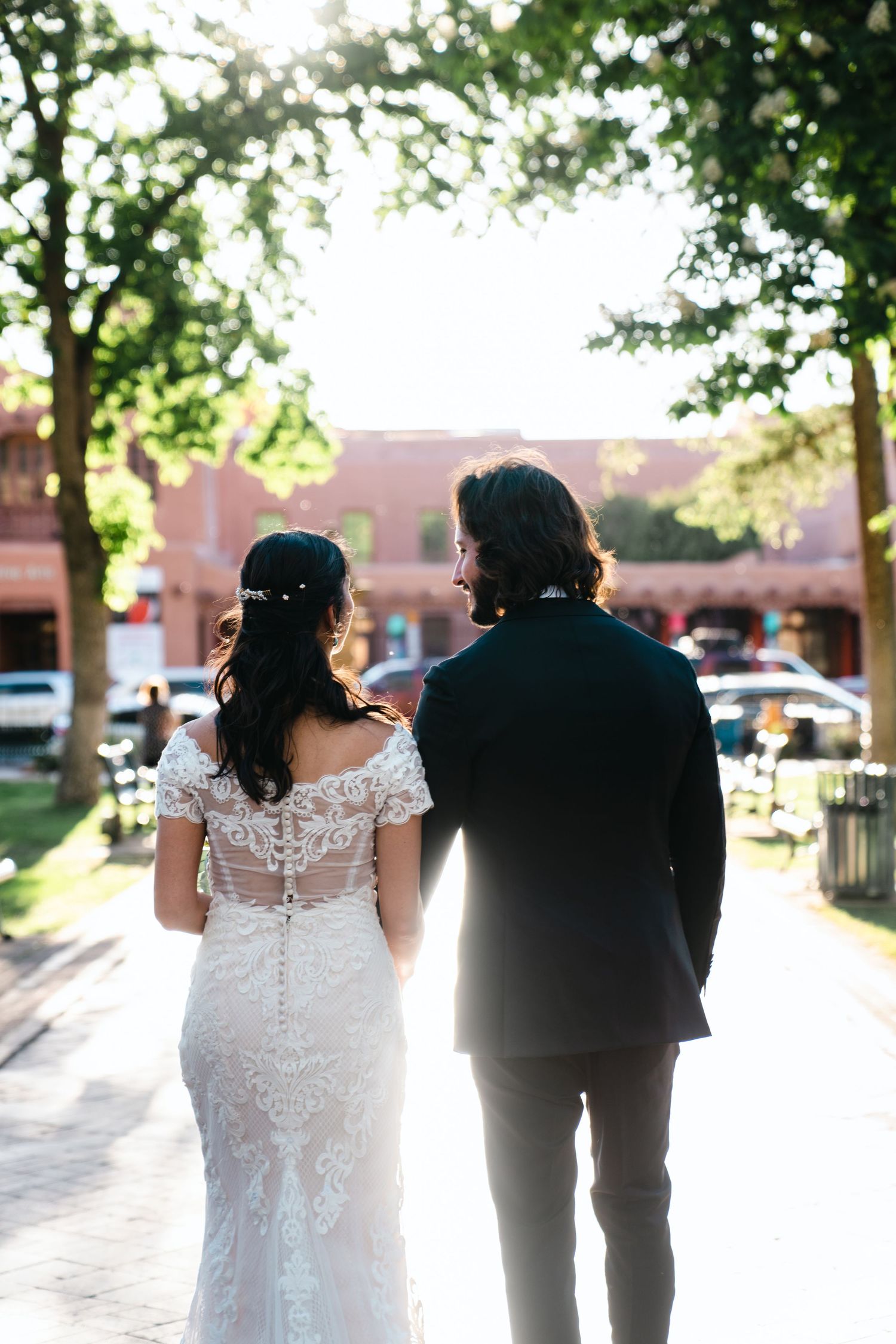 Wedding couple walking together in backlit sunlight through tree-lined path.