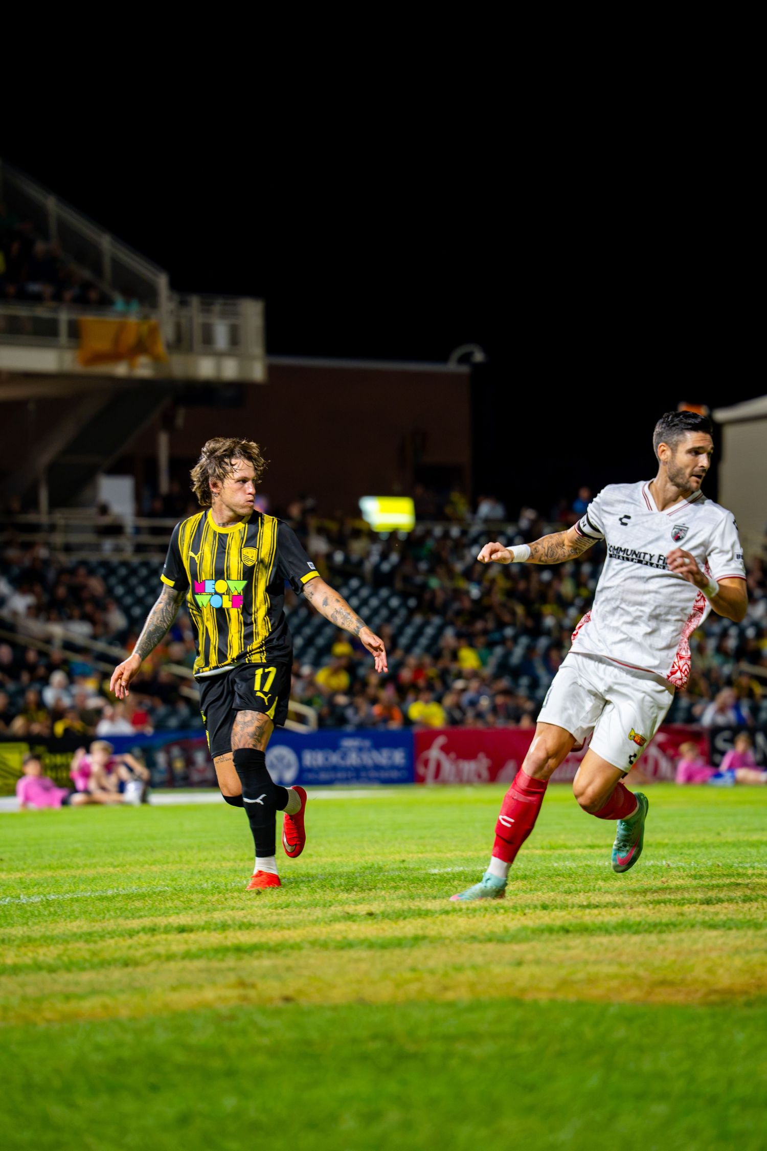 Two soccer players compete on a floodlit field, one in black and yellow, the other in white, during a night match.
