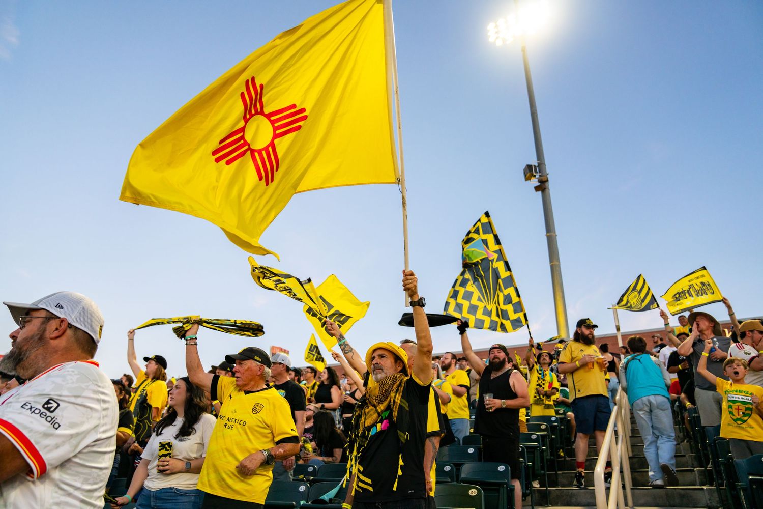 New Mexico United soccer fans wave yellow Zia flag and scarves in stadium stands during a sunny outdoor match.