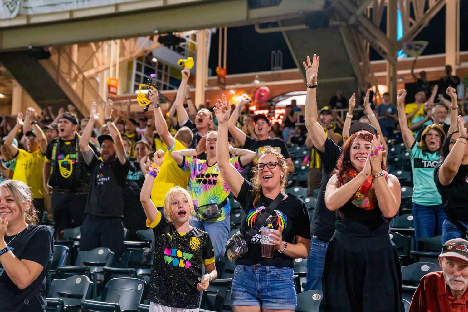 Excited sports fans cheering in stadium stands, wearing black and yellow team colors at a night game.