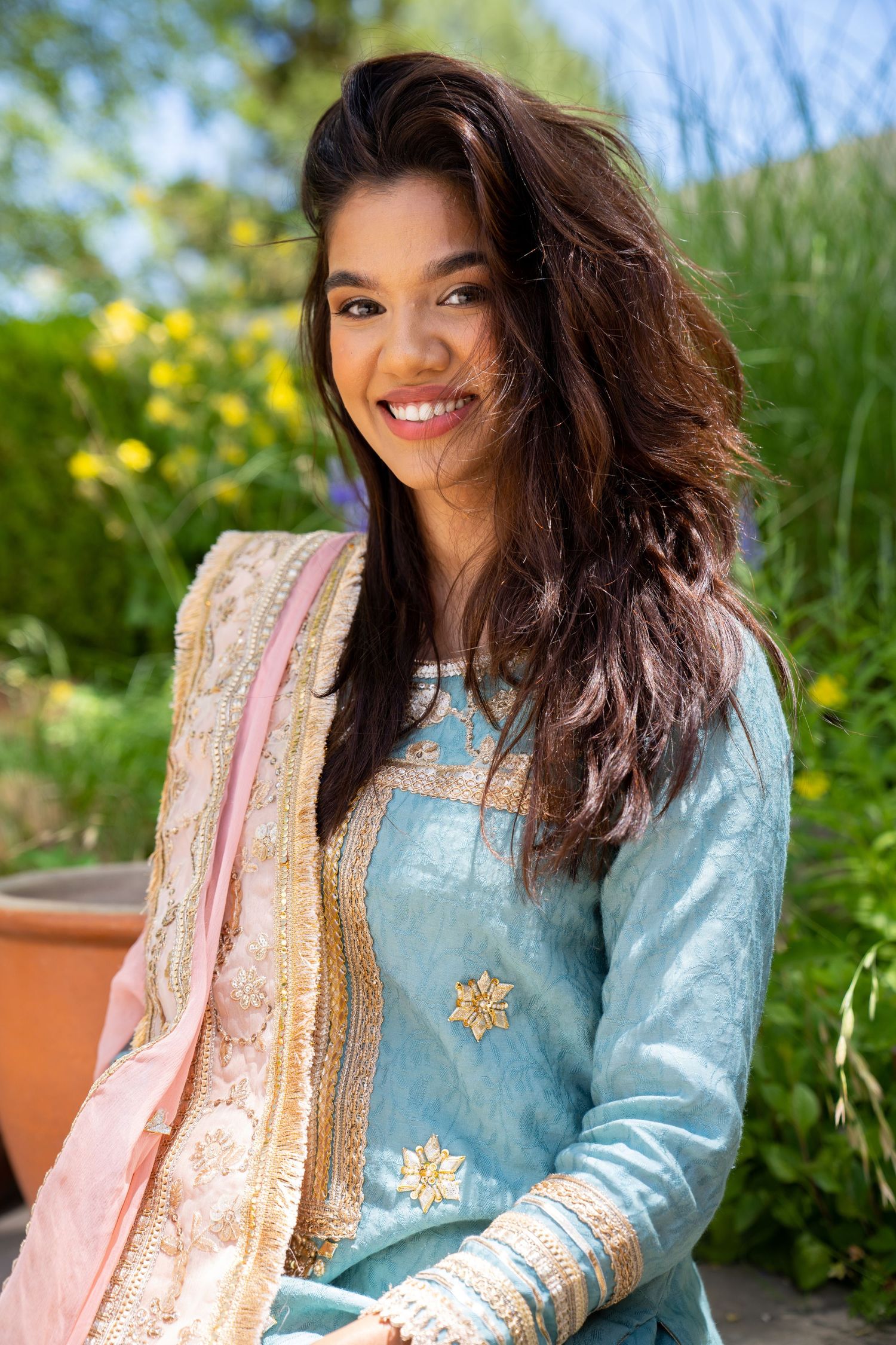 Portrait of person in traditional South Asian dress smiling outdoors with flowers in background.