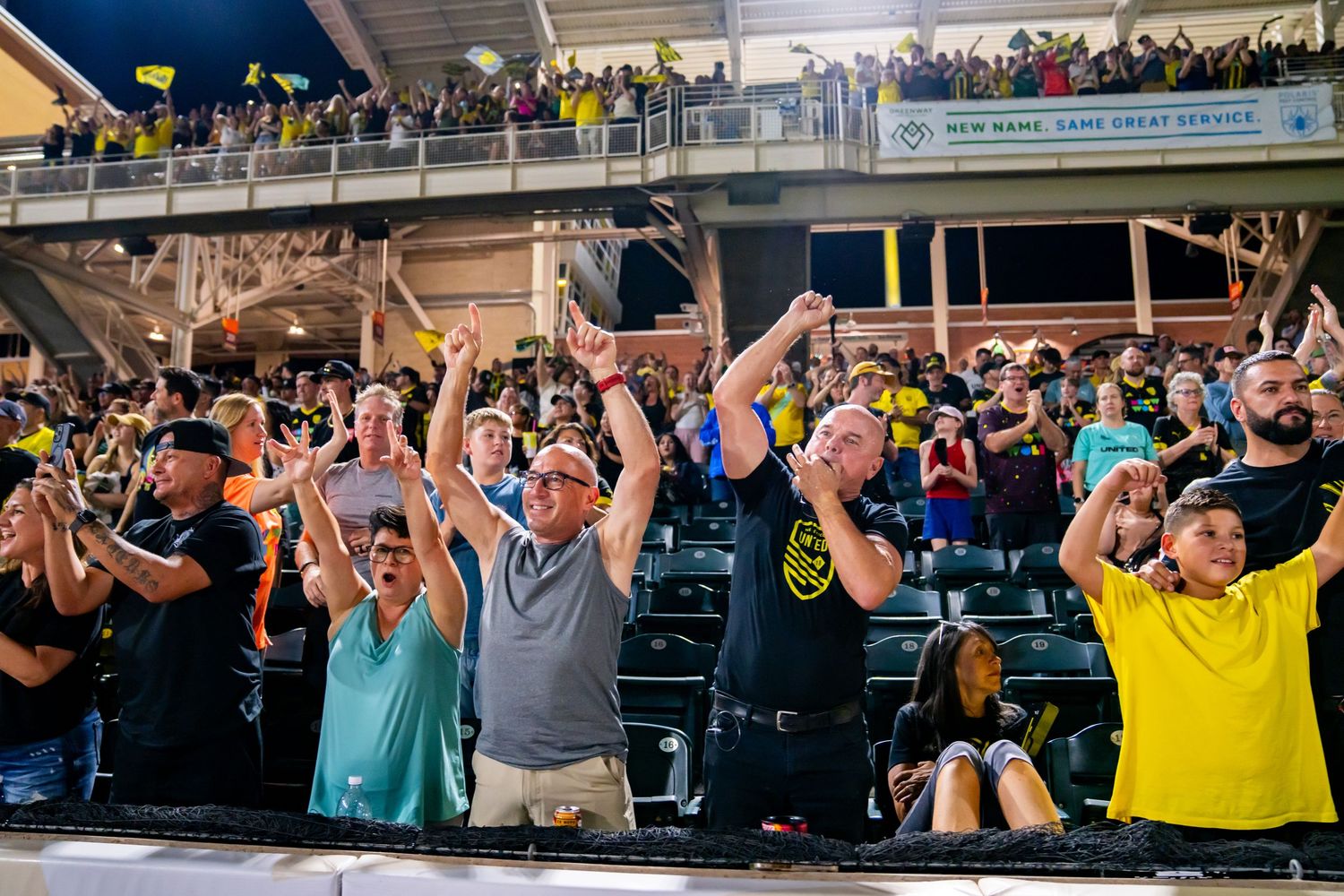 Excited soccer fans cheering in stadium stands wearing yellow and black team colors at a night game.