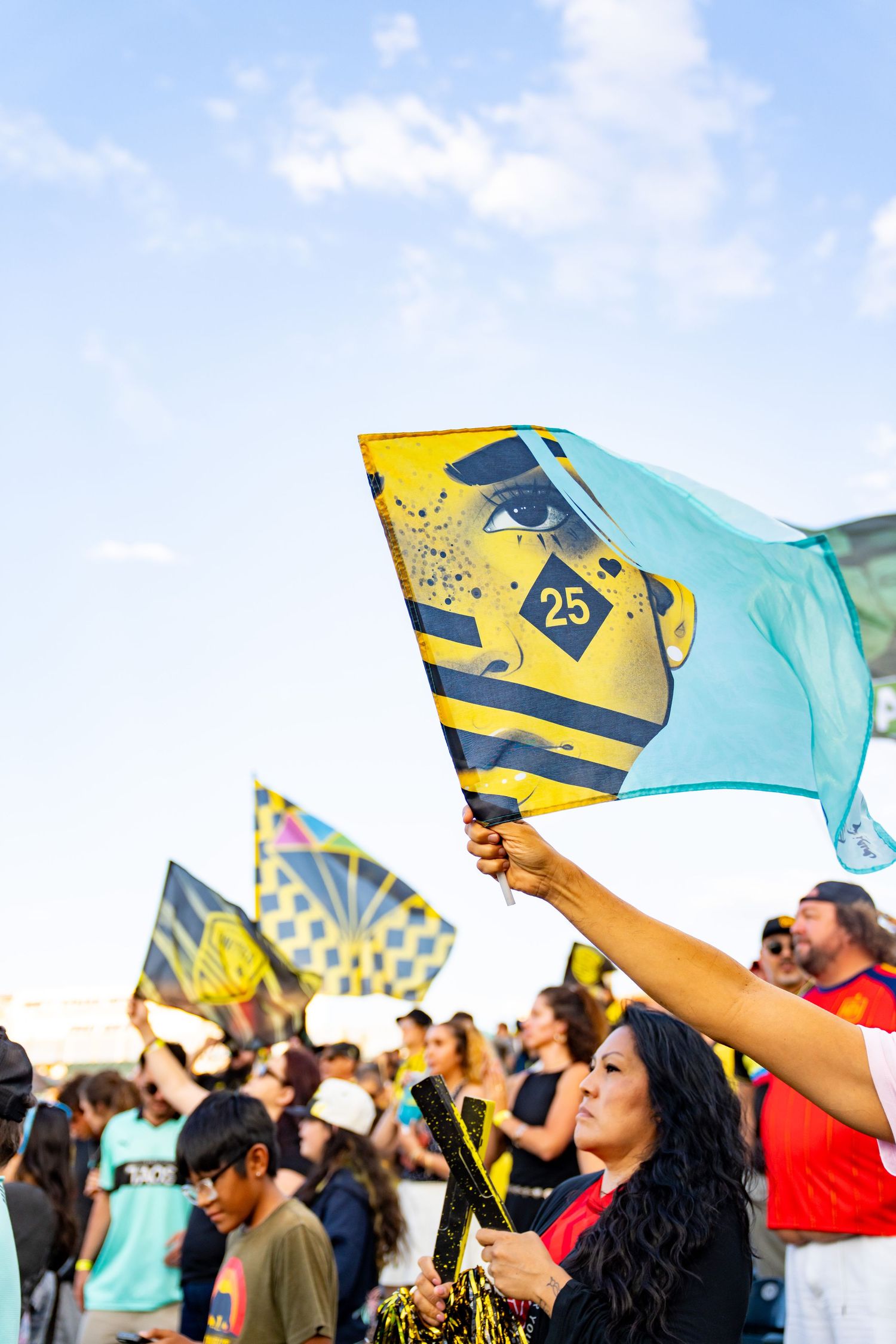 Woman waving yellow and blue flag numbered 25 at an outdoor sports rally with crowd and blue sky.