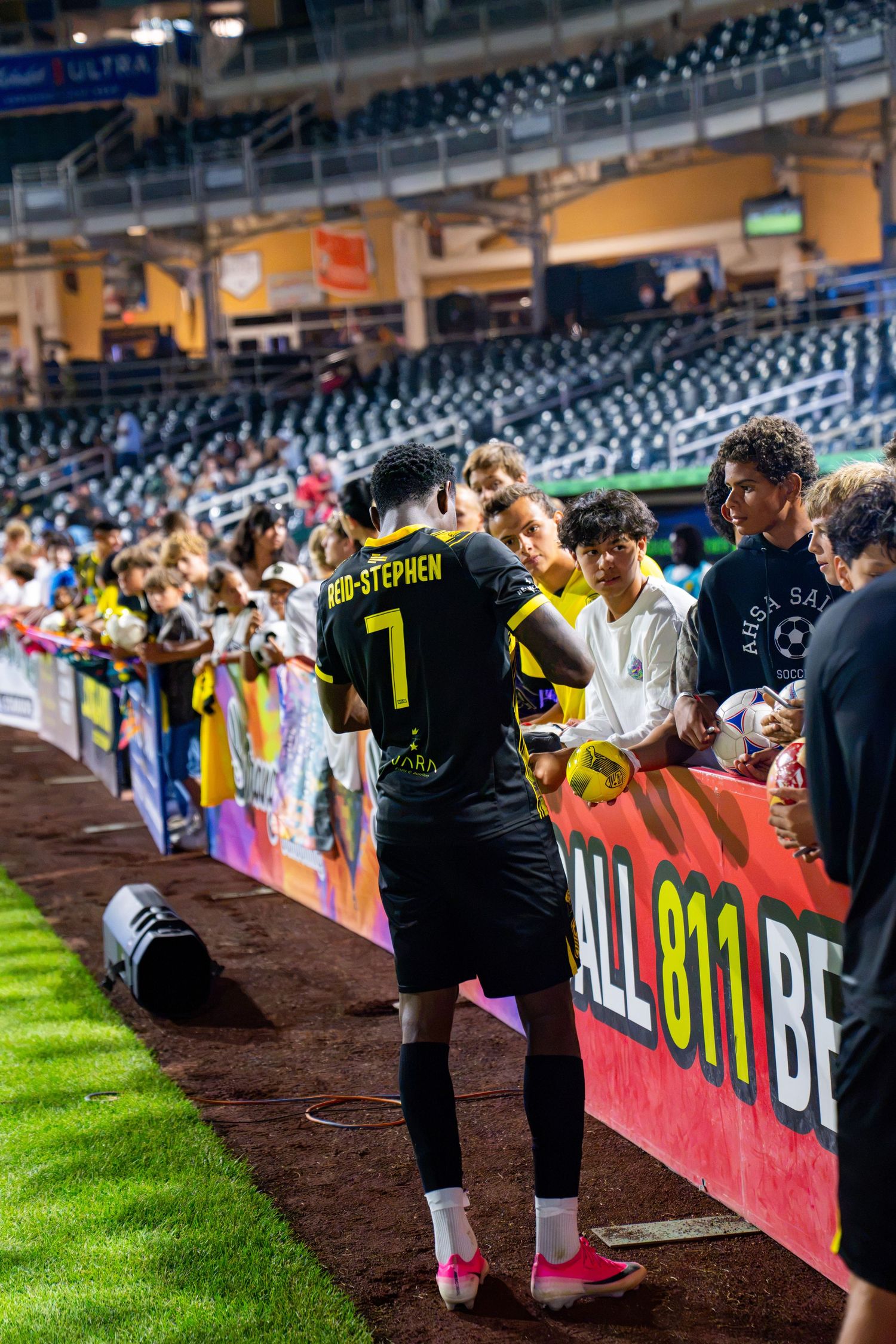 Soccer player #7 Rob-Stephen signs autographs for fans along the sideline barrier at a night game.