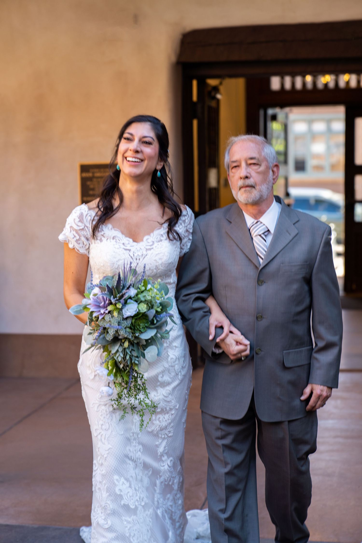 A wedding couple walking together in formal attire with the bride wearing a white lace dress and holding a bouquet.