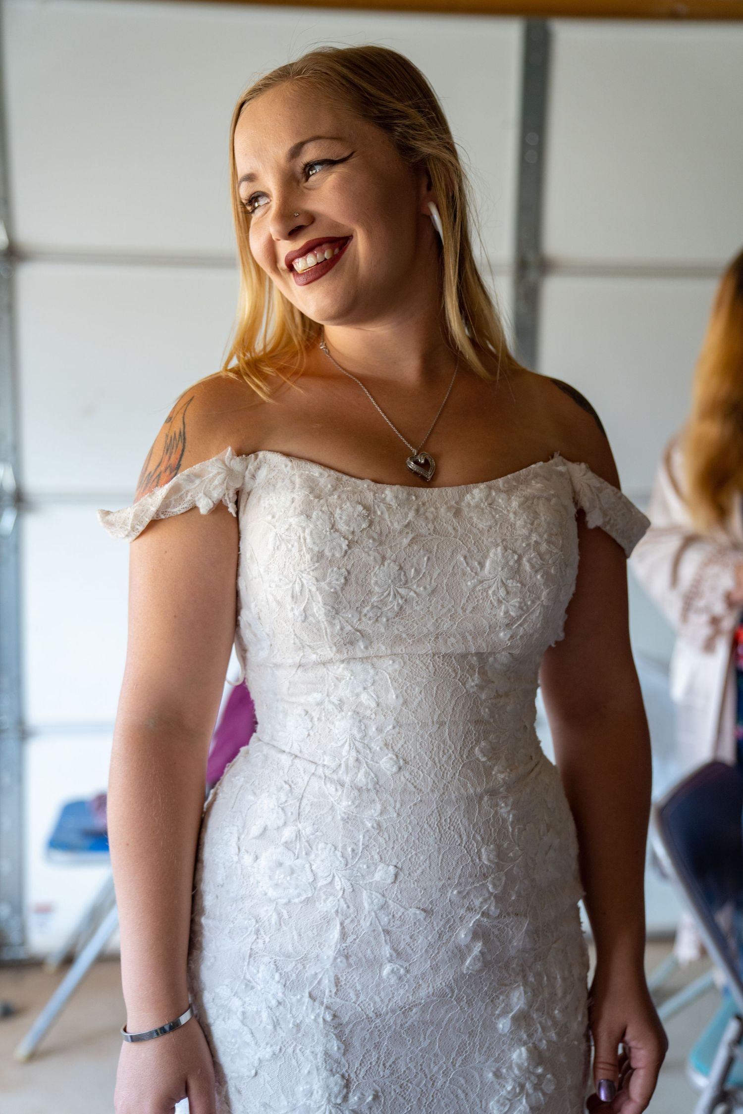 A bride wearing an off-shoulder white lace wedding dress smiles warmly while standing indoors.