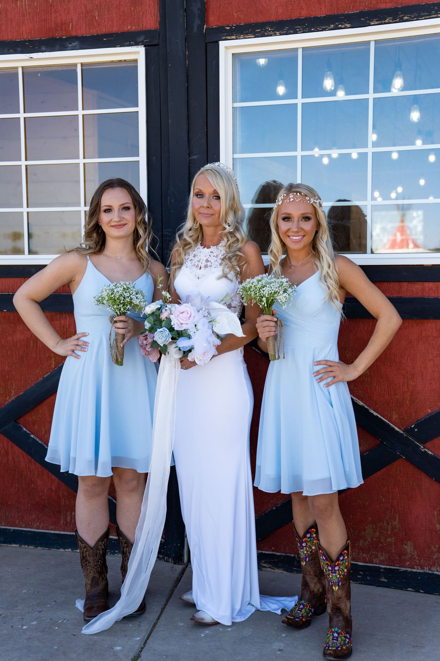 A group in light blue dresses and cowboy boots poses together against a red barn wall with string lights overhead.