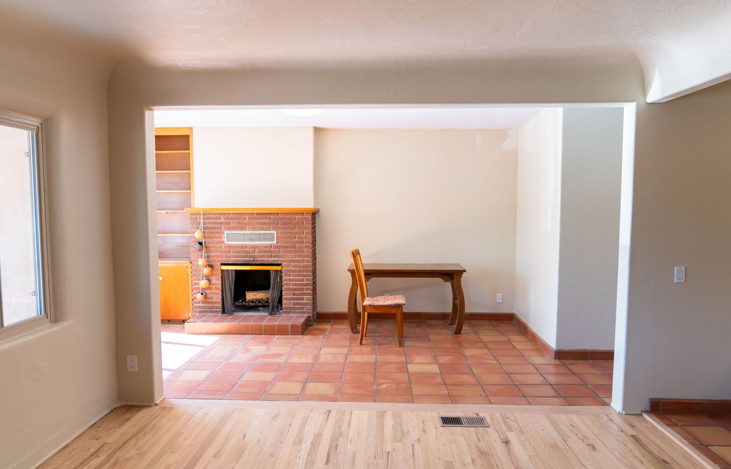Empty living room interior with hardwood floors, terracotta tiles, and a brick fireplace in the background.