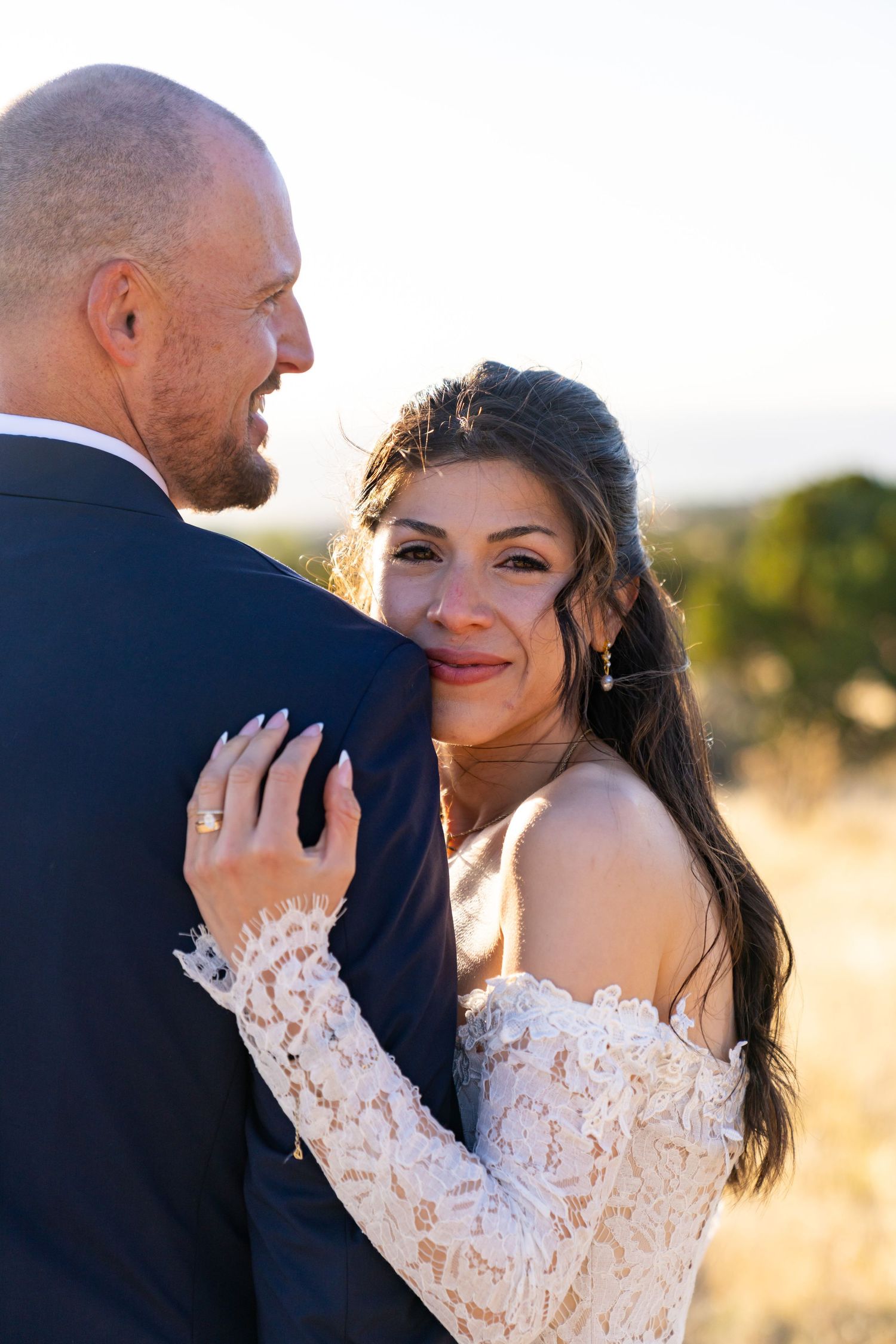 Bride in lace off-shoulder gown smiles at groom in navy suit during golden hour outdoor wedding portrait.