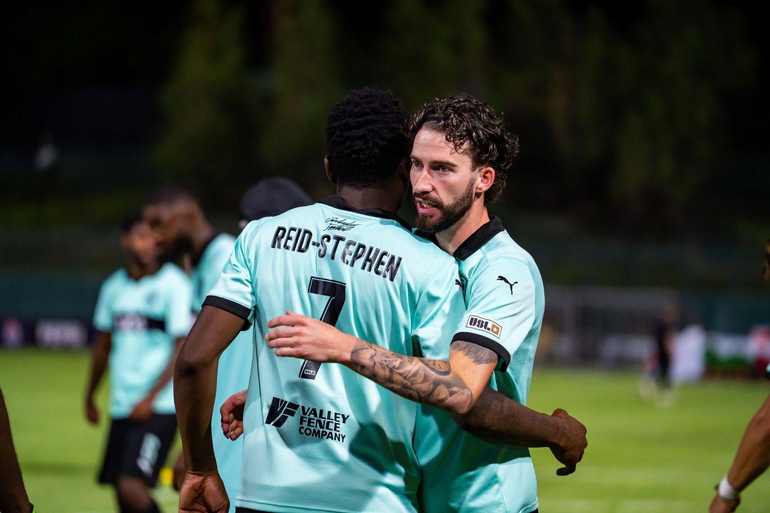 Two soccer players in teal uniforms embrace on field, with Reid-Stephen number 7 visible on back of jersey.