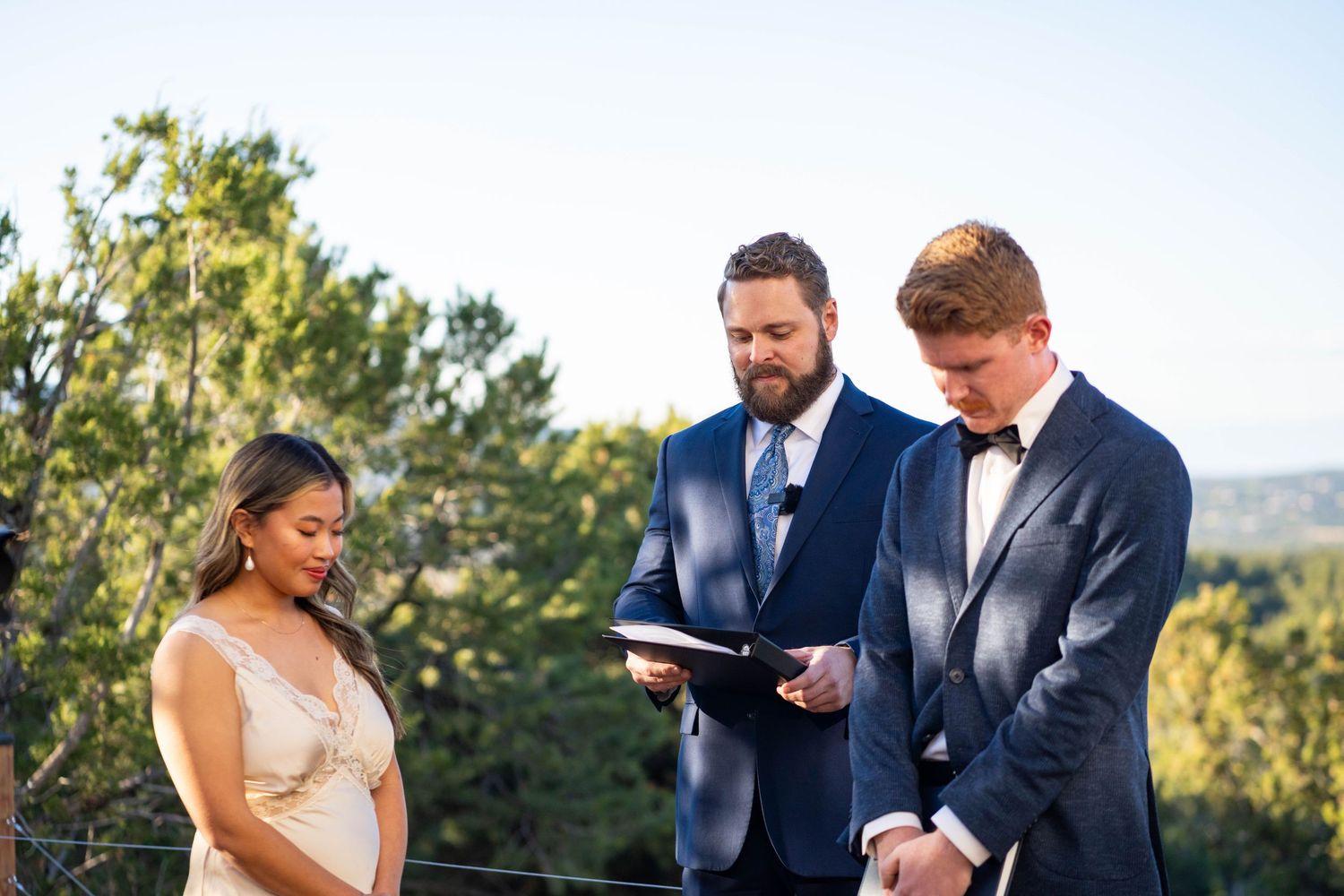 Wedding ceremony taking place outdoors with guests seated on white chairs among trees.