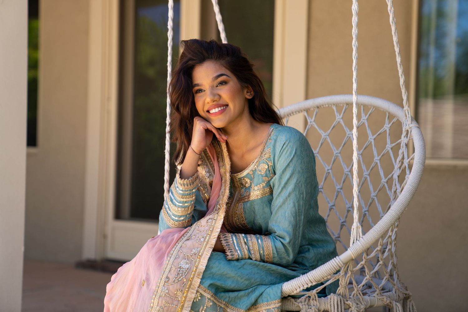 Person in traditional South Asian attire sitting in white hanging chair with joyful expression.