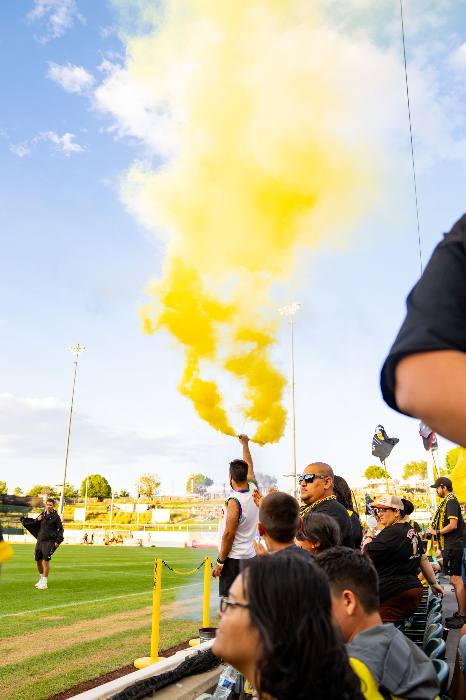 Fan holds yellow smoke flare at soccer stadium, crowds cheering in stands under blue sky.