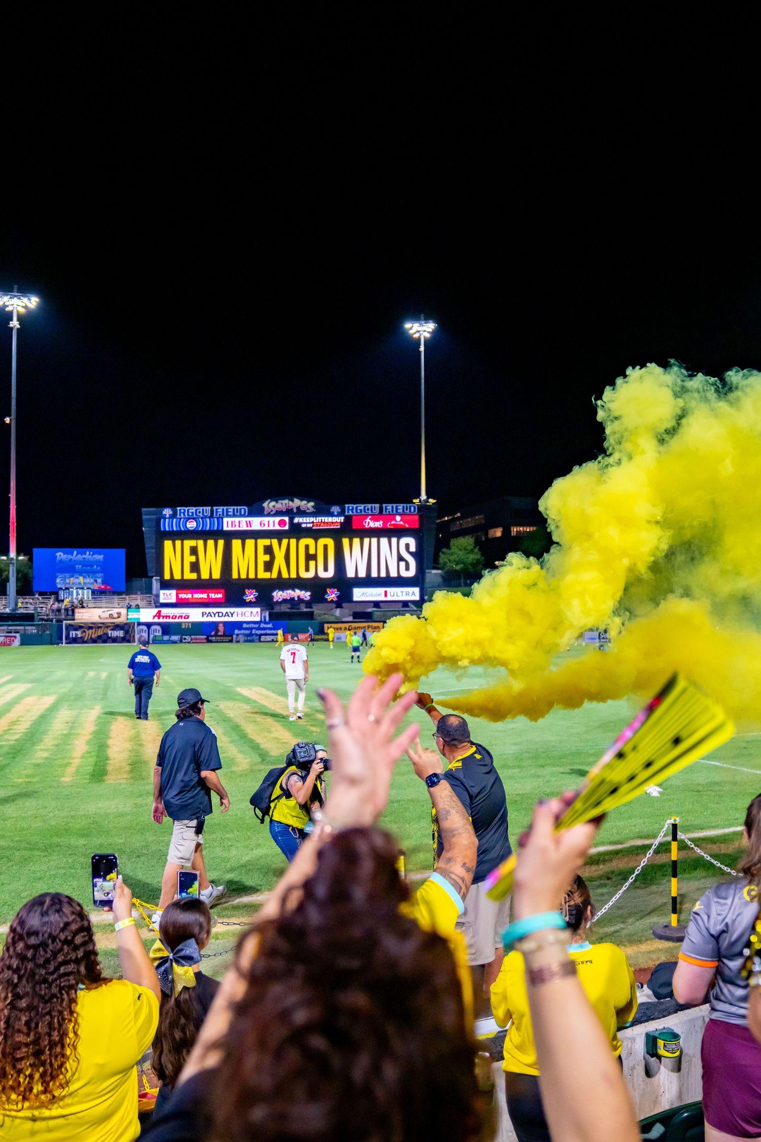 Fans celebrate with yellow smoke flares as the scoreboard reads New Mexico Wins at a night baseball game.