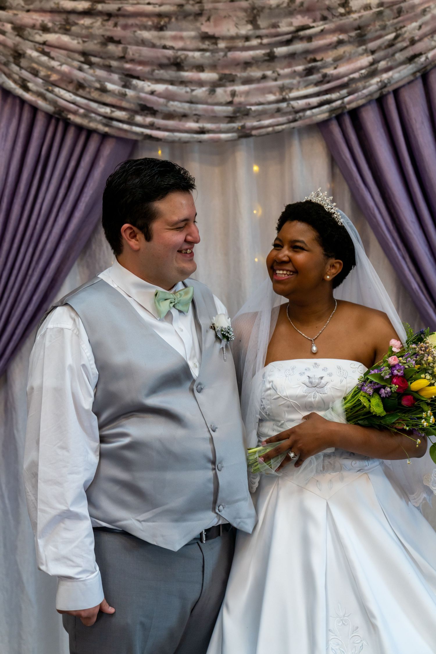 A joyful couple shares a happy moment at their wedding, with purple drapes and floral decor in the background.