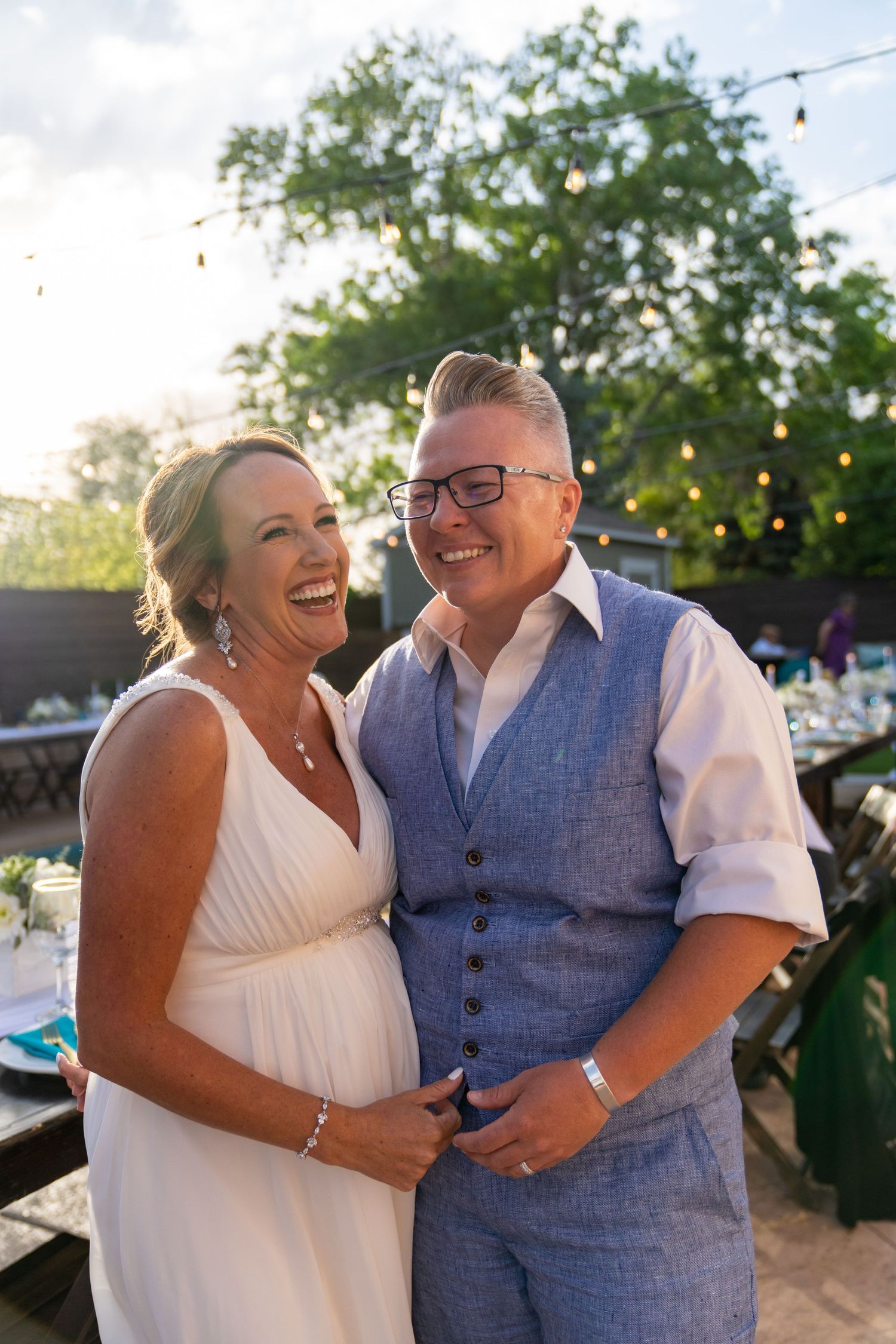 Happy couple shares joyful laughter during outdoor wedding celebration with string lights in background.