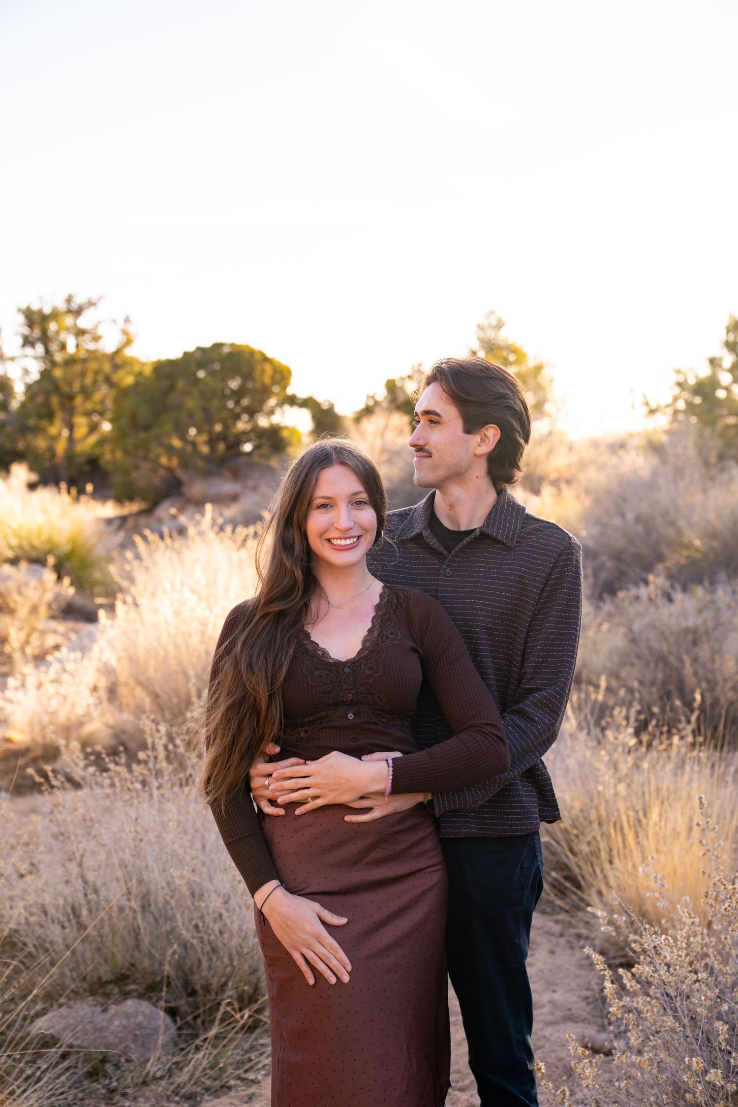 A couple poses outdoors at golden hour, man embracing woman from behind in desert landscape with shrubs and warm sunlight.
