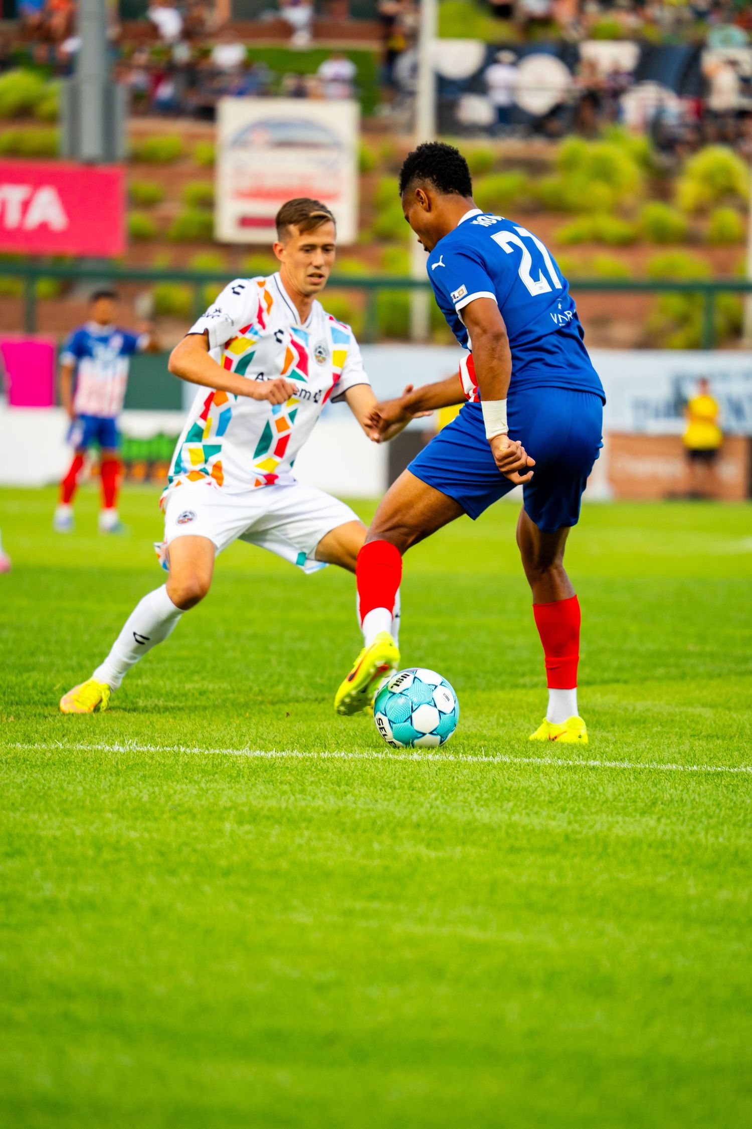 Two soccer players compete for the ball during a match, one in white and one in blue uniforms on a green field.