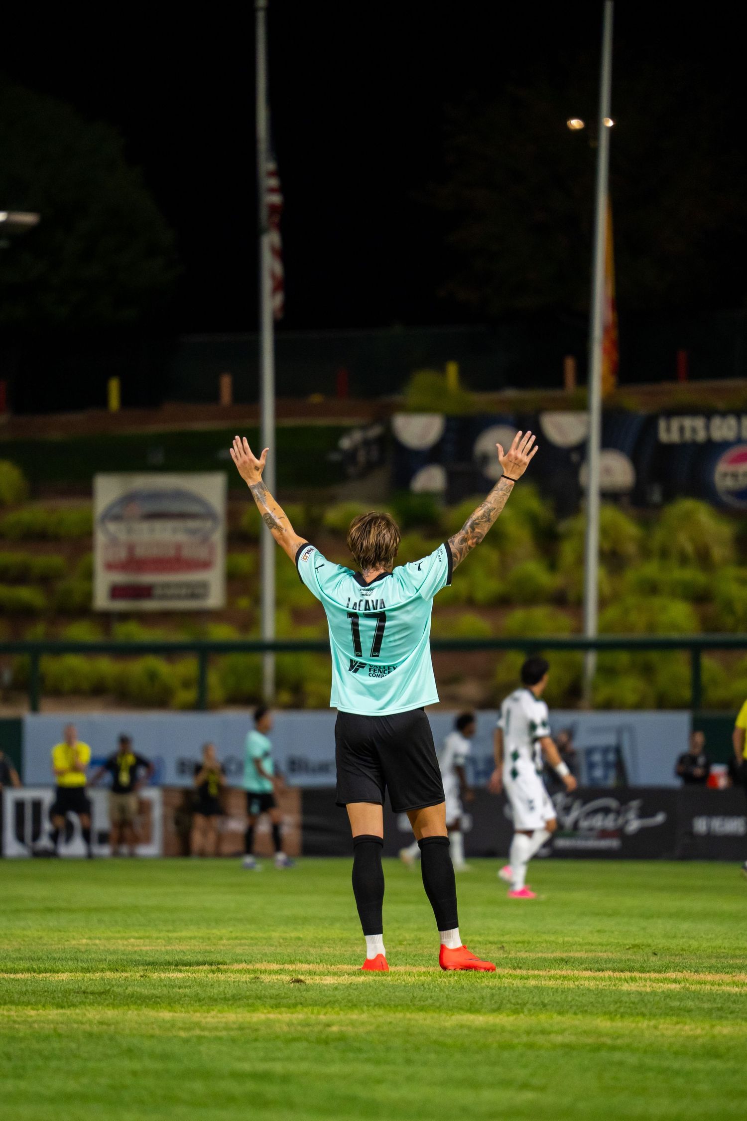 Soccer player #17 raises both arms on a night game field wearing a teal jersey with tattoos visible.