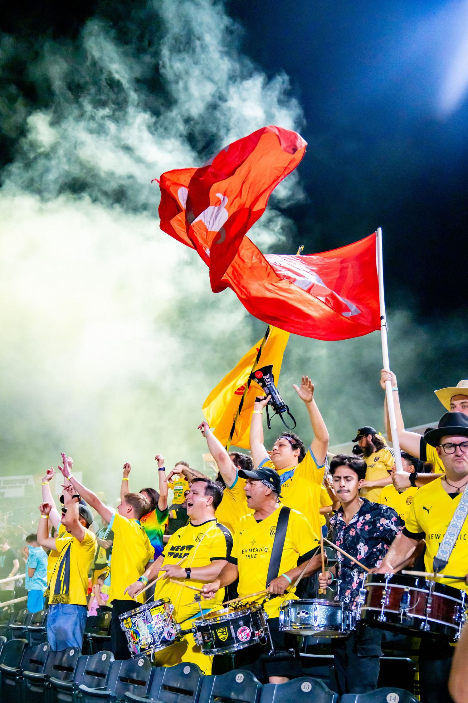 Enthusiastic soccer fans in yellow jerseys wave red flags and play drums in a stadium filled with colorful smoke.