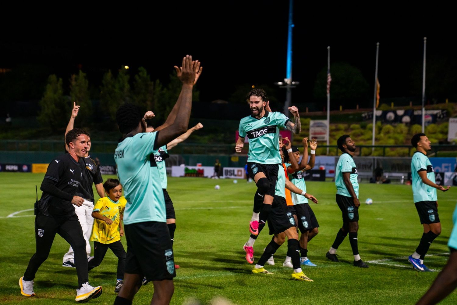 Soccer players celebrate on the field at night, high-fiving in teal jerseys under stadium lights.